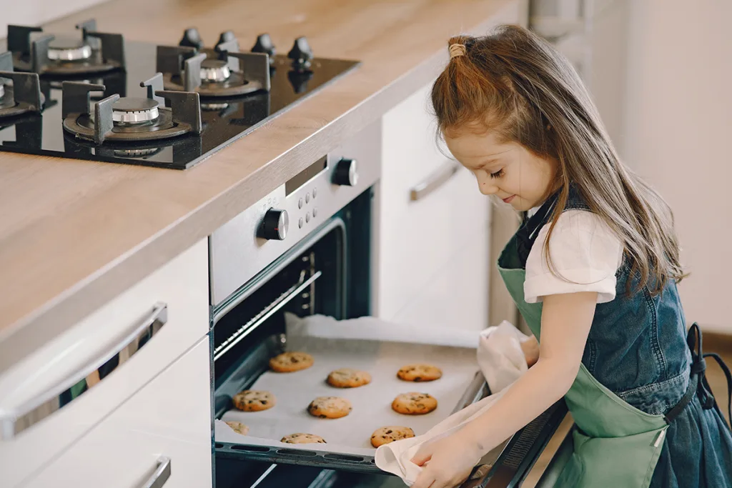 A young girl in an apron engages in American food culture as she pulls a baking pan of chocolate chip cookies from the oven.