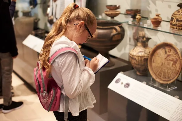 A girl sees ancient sculptures in a museum and intently takes notes, showing the value of teaching kids about other cultures.