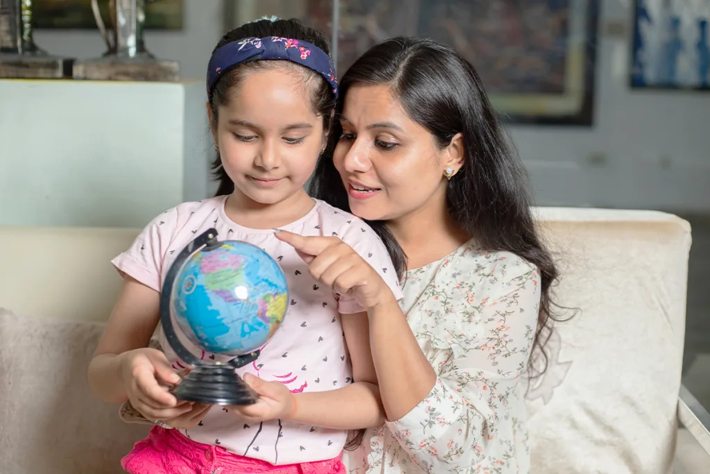 A young girl sits on her au pair's lap and holds a globe, which her au pair utilizes to compare Spain vs. Mexico culture.