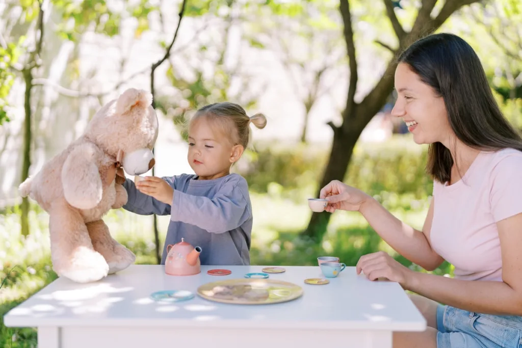 A woman smiles as a small girl serves her teddy bear at what might be the British tradition of afternoon tea outside.