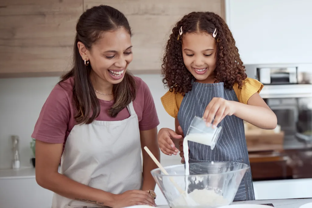 A smiling au pair and a young girl bonding while baking together in the kitchen, pouring ingredients into a mixing bowl.