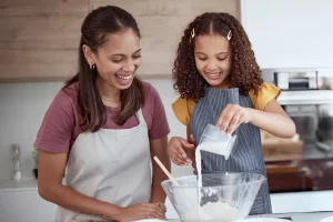 A smiling au pair and a young girl bonding while baking together in the kitchen, pouring ingredients into a mixing bowl.